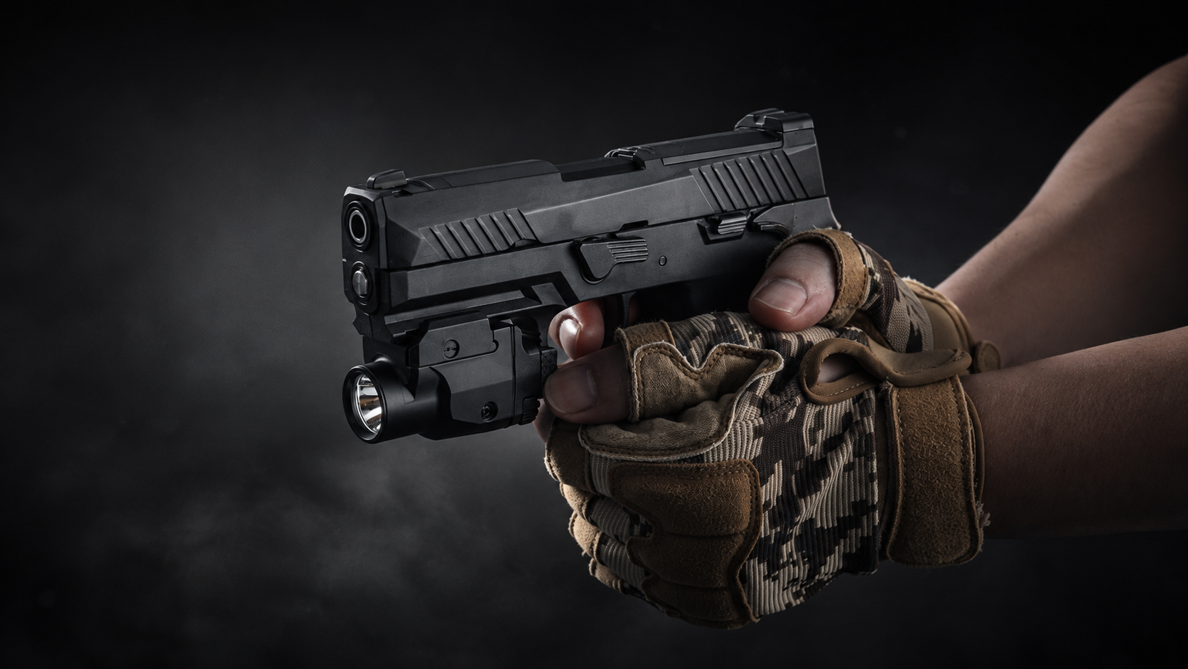 A black tactical pistol with an attached flashlight, held by a person wearing tan and brown camouflage fingerless gloves against a dark smoky background.