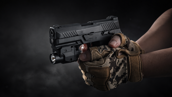A black tactical pistol with an attached flashlight, held by a person wearing tan and brown camouflage fingerless gloves against a dark smoky background.