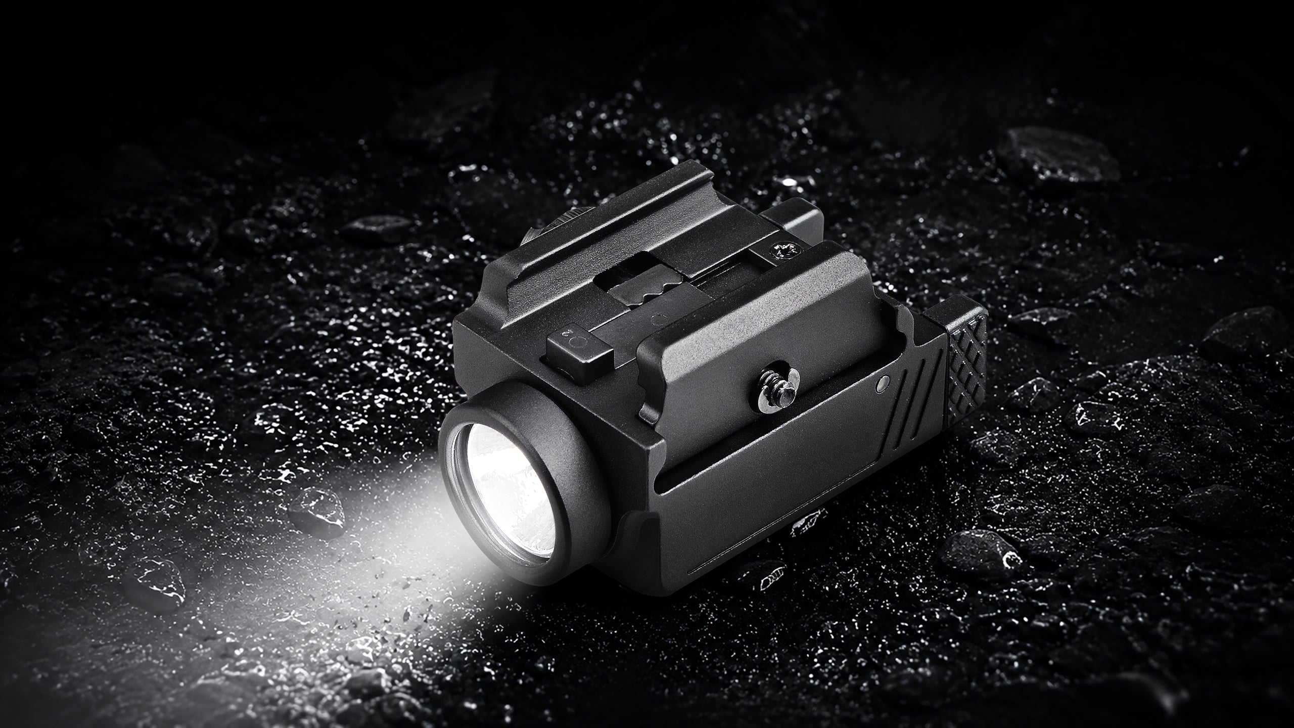 A tactical black flashlight with a bright illuminated lens, shown resting on a dark, wet, rocky surface. The flashlight has a mounting bracket on top and is casting a bright beam of light to the left.