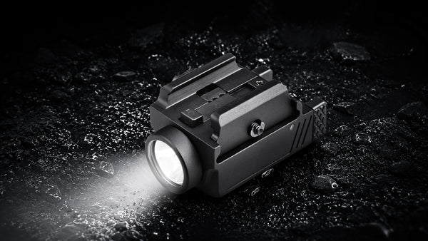 A tactical black flashlight with a bright illuminated lens, shown resting on a dark, wet, rocky surface. The flashlight has a mounting bracket on top and is casting a bright beam of light to the left.